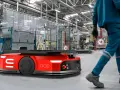 Czerwone roboty AGV poruszają się po hali produkcyjnej, gdzie pracownicy w niebieskich kombinezonach nadzorują procesy.