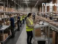 Kobieta w kamizelce odblaskowej nadzoruje pracę magazynu, gdzie trwa automatyzacja logistyki. Pracownicy sortują paczki na taśmie.