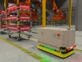 Wózki AGV w magazynie, obok regałów z czerwonymi pojemnikami, przygotowane do transportu.