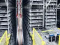 Wysokie regały wypełnione pojemnikami, z widocznymi elementami automatyzacji magazynu, takimi jak przenośniki i czujniki.