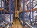 Automatyczny **stacker crane** porusza się po torach w magazynie wysokiego składowania, między regałami pełnymi towarów.