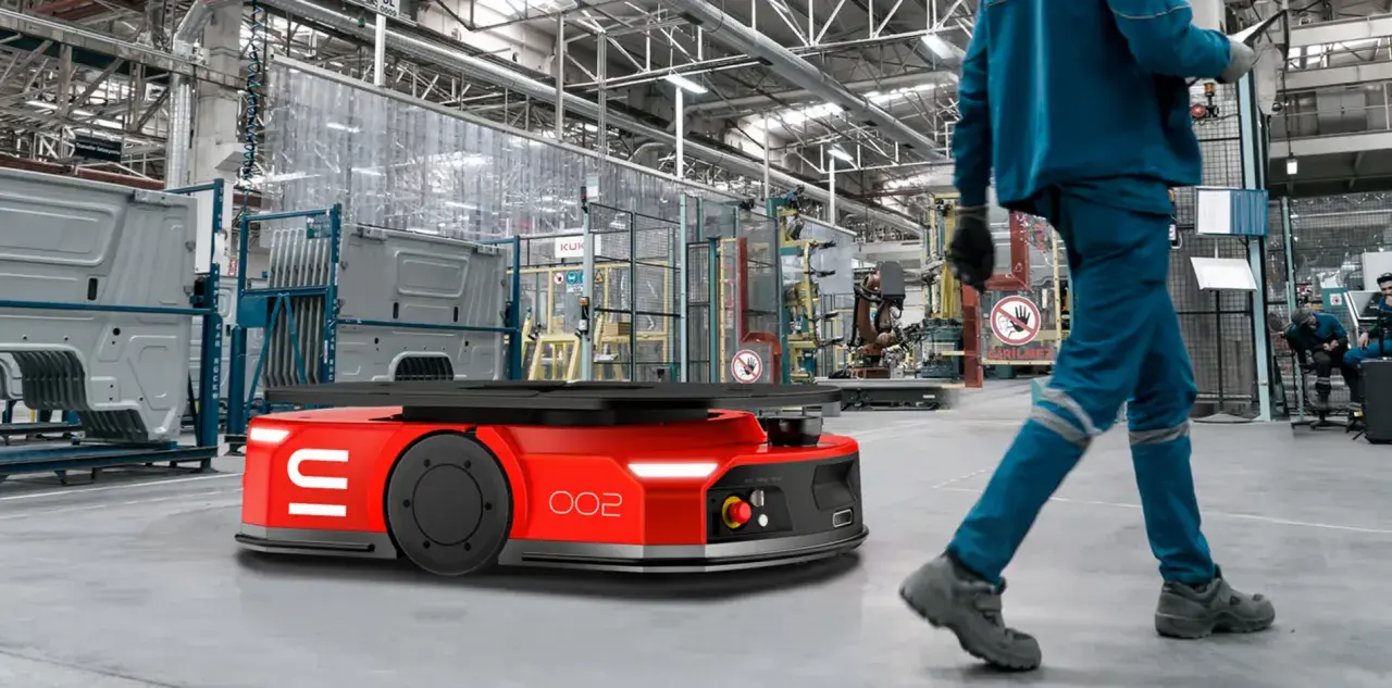 Czerwone roboty AGV poruszają się po hali produkcyjnej, gdzie pracownicy w niebieskich kombinezonach nadzorują procesy.