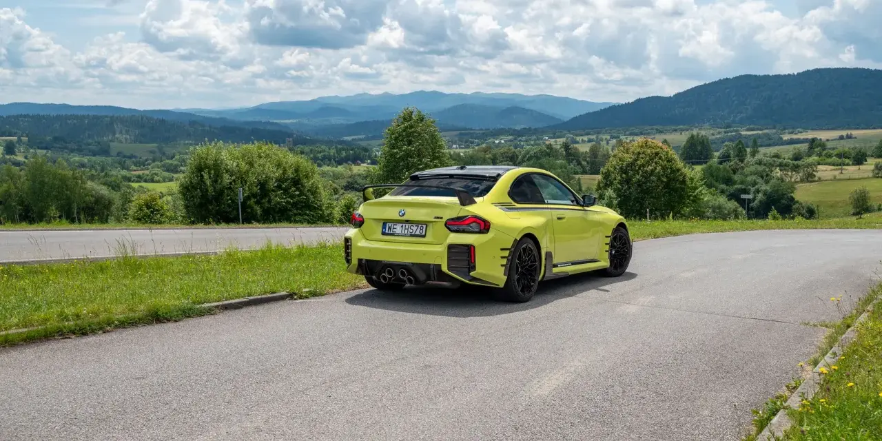 Żółte BMW M2 z czterema końcówkami wydechu, czyli otwór pod M4, na tle malowniczych gór i chmur.