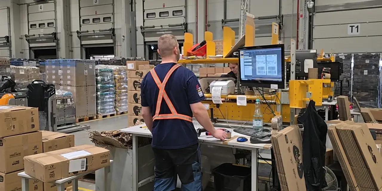 Pracownik w magazynie produkcyjnym przy stanowisku komputerowym, otoczony paletami z towarami i bramami dokowymi.