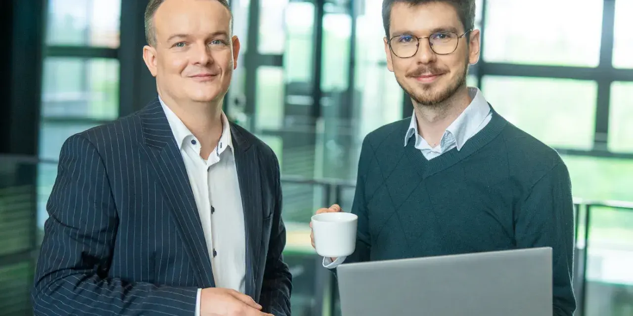 Dwóch mężczyzn w biurze, jeden z laptopem, drugi z kawą. Pracują nad projektem w systemie Symfonia.