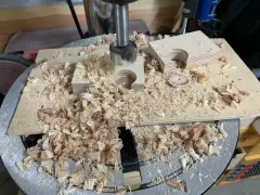 Wiertło pogłębia otwory w sklejce, tworząc elementy do budowy frezarki. Wióry drewna pokrywają blat roboczy.