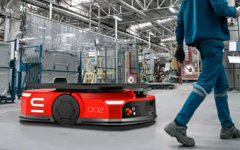 Czerwone roboty AGV poruszają się po hali produkcyjnej, gdzie pracownicy w niebieskich kombinezonach nadzorują procesy.