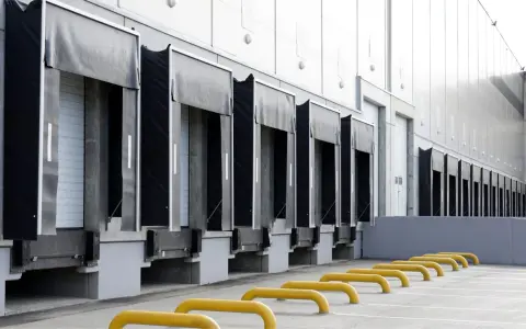 Rząd doków załadunkowych z czarnymi fartuchami i białymi drzwiami. Żółte bariery parkingowe na pierwszym planie.