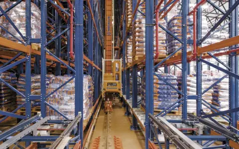 Automatyczny **stacker crane** porusza się po torach w magazynie wysokiego składowania, między regałami pełnymi towarów.
