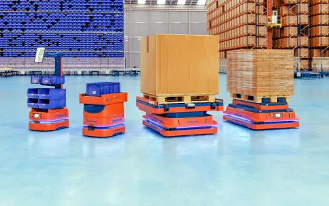 Pomarańczowe roboty AMR, czyli zautomatyzowane wozy magazynowe, przewożą paczki i pojemniki w nowoczesnym magazynie.