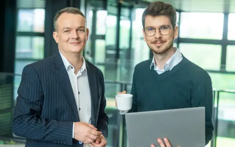 Dwóch mężczyzn w biurze, jeden z laptopem, drugi z kawą. Pracują nad projektem w systemie Symfonia.