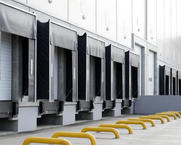 Rząd doków załadunkowych z czarnymi fartuchami i białymi drzwiami. Żółte bariery parkingowe na pierwszym planie.