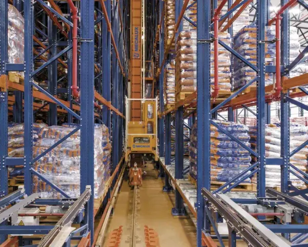 Automatyczny **stacker crane** porusza się po torach w magazynie wysokiego składowania, między regałami pełnymi towarów.