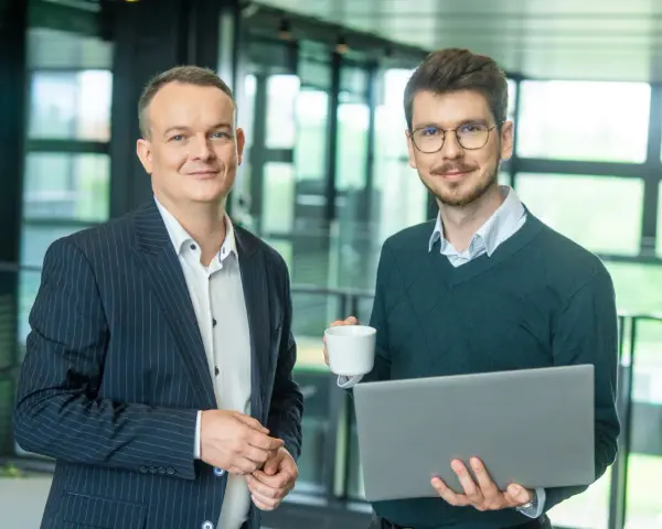Dwóch mężczyzn w biurze, jeden z laptopem, drugi z kawą. Pracują nad projektem w systemie Symfonia.