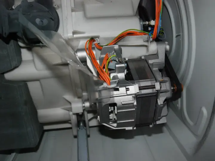 Silnik pralki z paskiem napędowym i przewodami. Widać elementy mechaniczne, które wpływają na wzór na prędkość obrotową bębna.