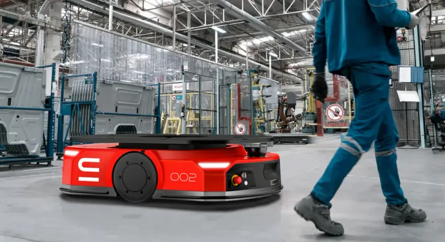 Czerwone roboty AGV poruszają się po hali produkcyjnej, gdzie pracownicy w niebieskich kombinezonach nadzorują procesy.
