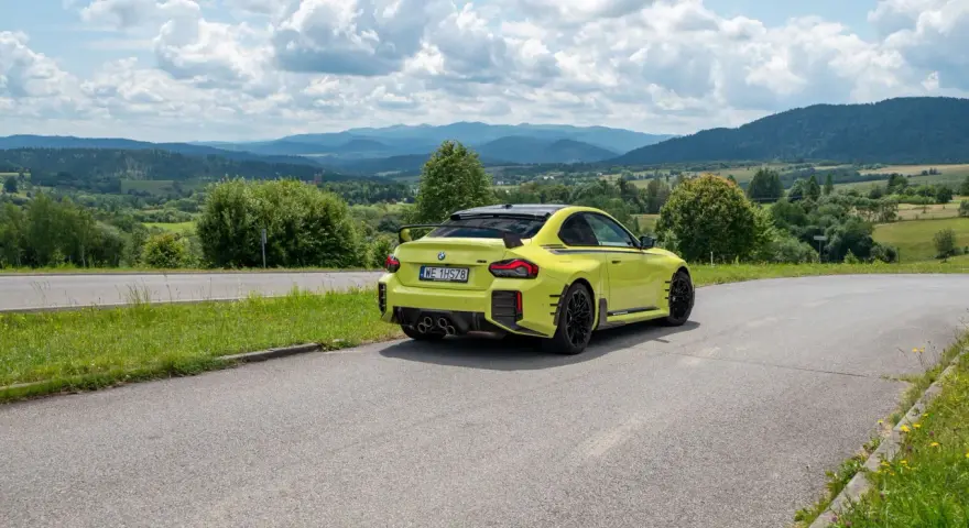 Żółte BMW M2 z czterema końcówkami wydechu, czyli otwór pod M4, na tle malowniczych gór i chmur.