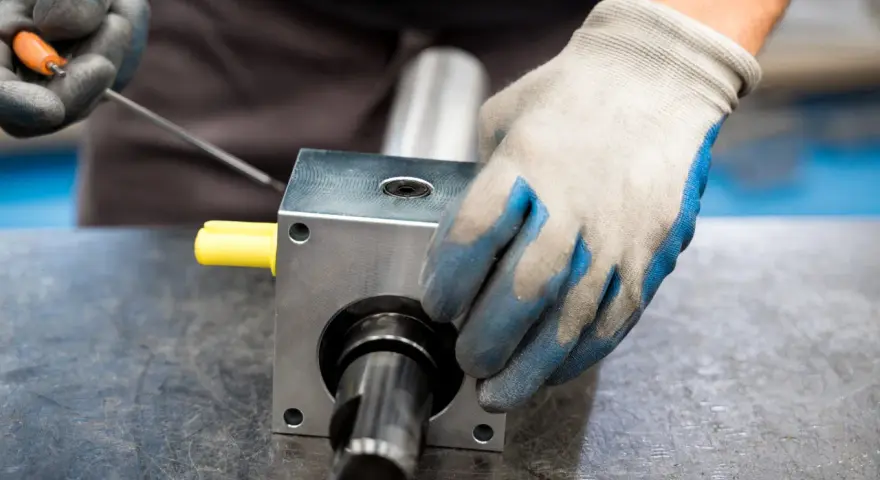 Ręce w rękawiczkach montują element mechaniczny, prawdopodobnie sprzęgła zębate, używając narzędzi.