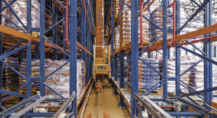 Automatyczny **stacker crane** porusza się po torach w magazynie wysokiego składowania, między regałami pełnymi towarów.