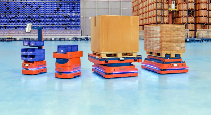 Pomarańczowe roboty AMR, czyli zautomatyzowane wozy magazynowe, przewożą paczki i pojemniki w nowoczesnym magazynie.