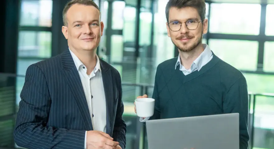 Dwóch mężczyzn w biurze, jeden z laptopem, drugi z kawą. Pracują nad projektem w systemie Symfonia.