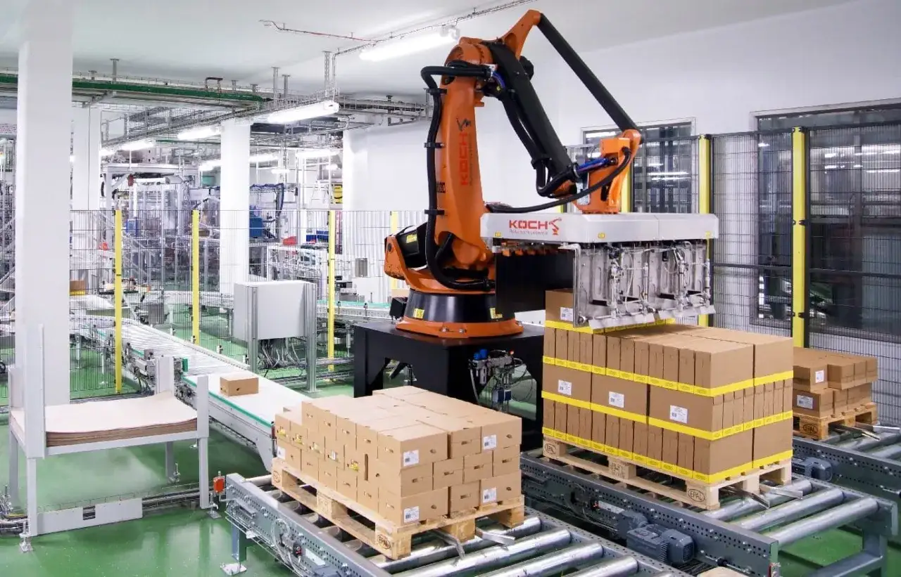 Pomarańczowy robot przemysłowy z chwytakiem pracuje nad układaniem kartonów na paletach. To zaawansowane systemy paletyzacji w akcji.