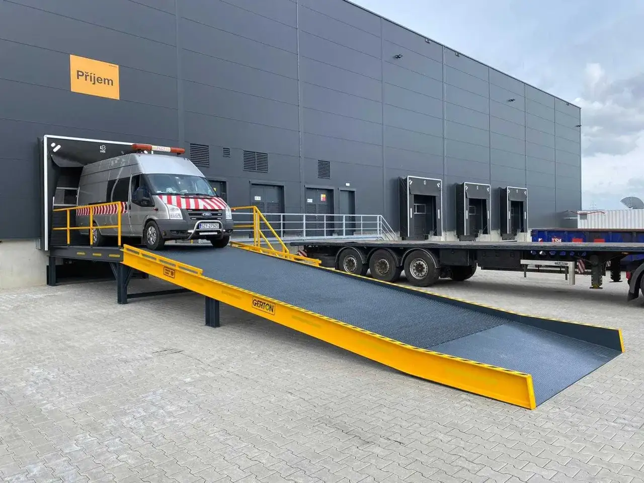 Van wjeżdża na rampę przy nowoczesnym loading dock. Obok stoi przyczepa ciężarówki.